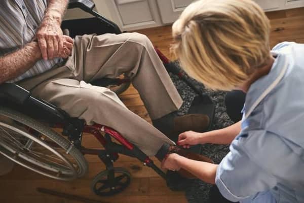 Female healthcare worker helping disabled senior man in wheelchair, tie shoe laces. Credit: Getty Images