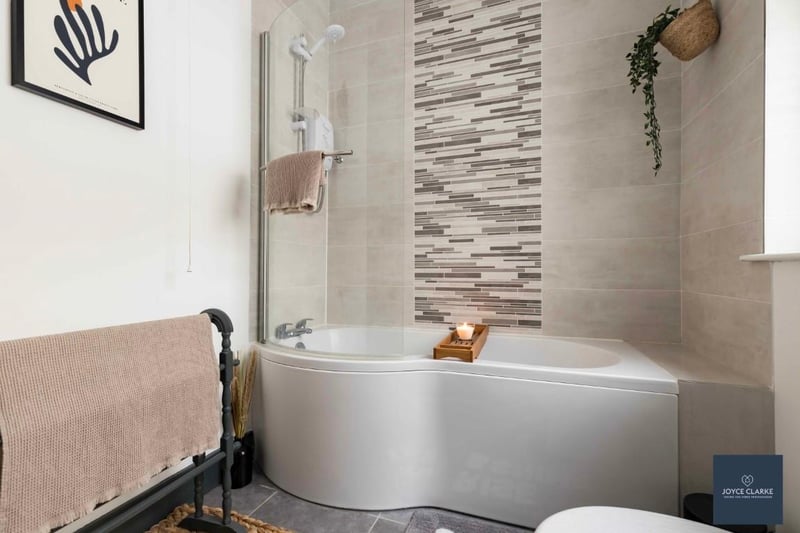 The modern bathroom has a P-shaped bath with Triton power shower, a dual flush WC, sink with vanity unit and a heated towel rail. There is a tiled floor and part tiled wall.