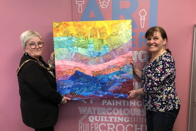 Mid and East Antrim Deputy Mayor, Cllr Beth Adger, standing with Ann McKeown showing off the first sensory art painting done a few years ago which has a permanent place hanging on the wall in Ballymena Central Library